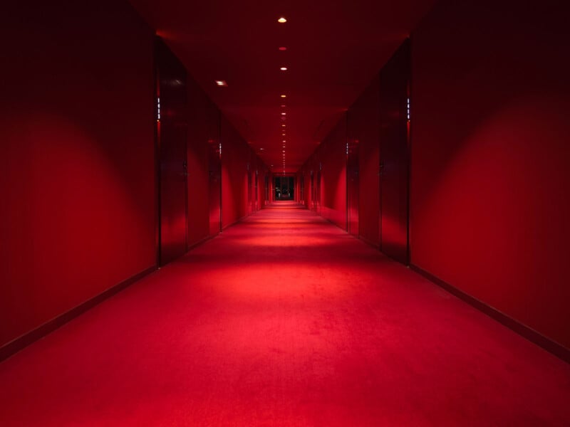A long, red-carpeted hallway with red walls and dim overhead lighting. The perspective leads to a dark doorway at the far end, creating a dramatic and moody atmosphere.