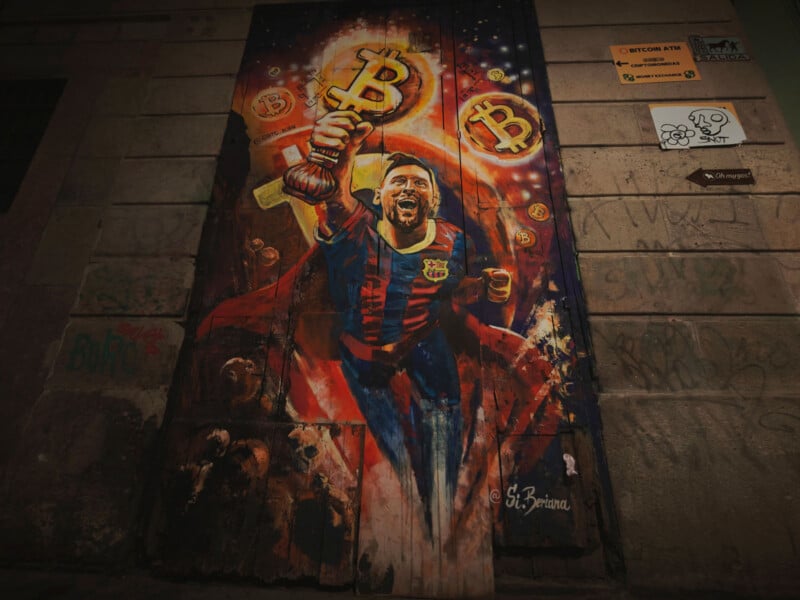 A vibrant mural on a brick wall depicts a jubilant soccer player in a red and blue uniform, holding a golden Bitcoin trophy. Cryptocurrency symbols surround him. Graffiti and signage are present on the wall.