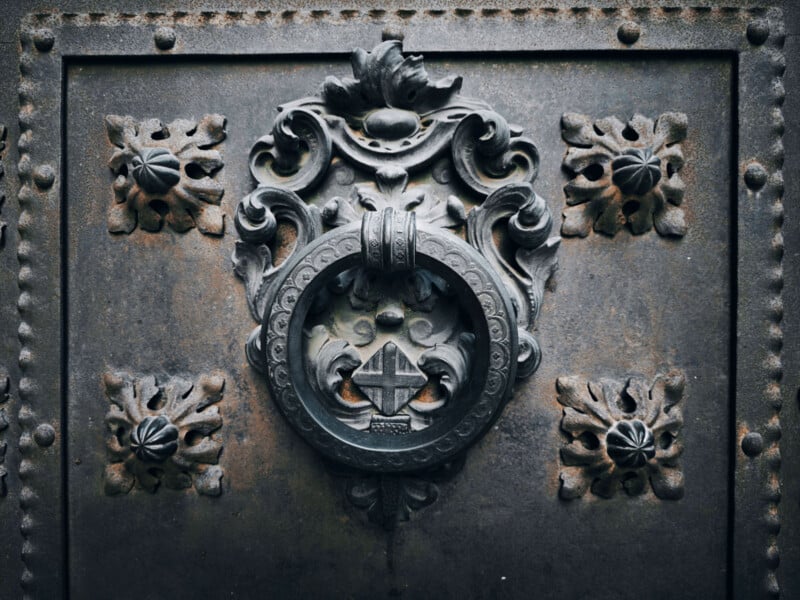 An ornate metal door knocker with intricate floral and geometric designs on an aged, textured surface. The central ring features a square pattern inside, surrounded by decorative motifs.